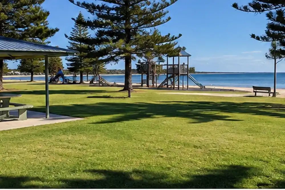 Grassy seaside reserve with shaded picnic tables, playground and calm blue water